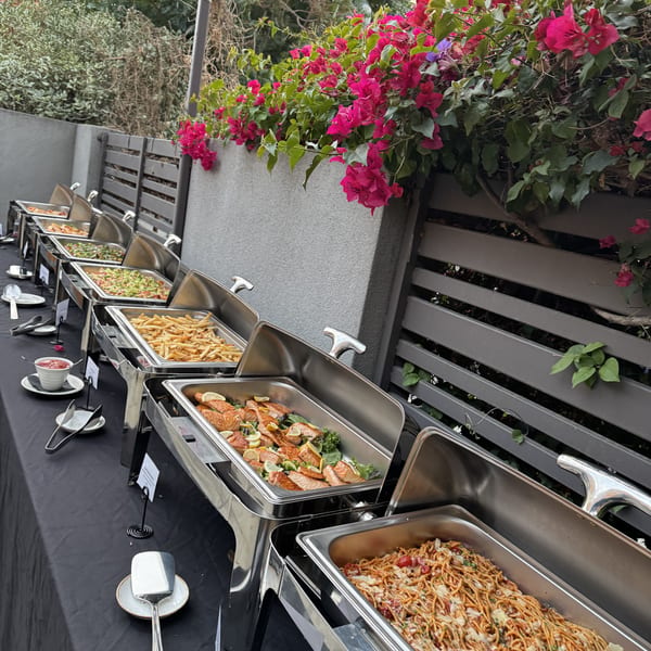 Outdoor catering buffet featuring chafing dishes with pasta, salmon, fries, and salads next to pink flowers.