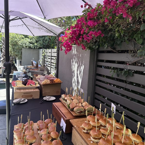 Outdoor buffet table with beef sliders, chicken sliders, sandwiches, charcuterie, and fruit under white umbrellas.