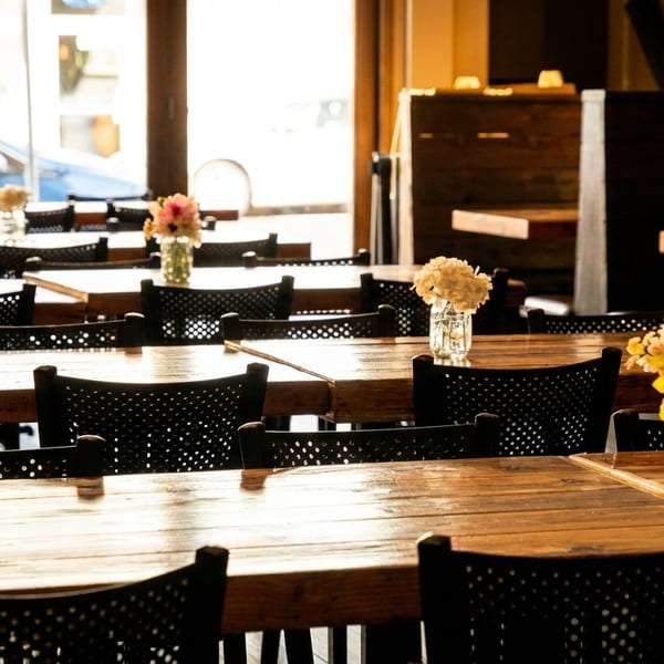 Empty restaurant dining room with wooden tables, black chairs, and small flower arrangements.