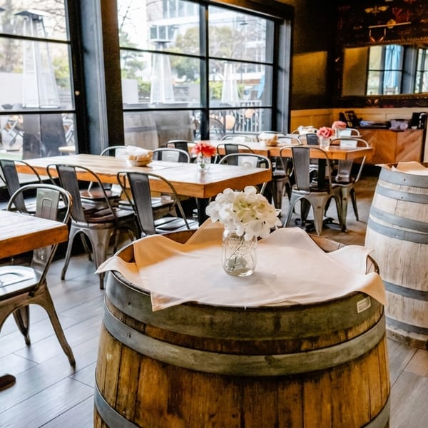 Restaurant interior with wooden tables, metal chairs, wine barrels, and large windows overlooking a patio.