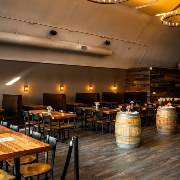 Spacious restaurant interior with long wooden tables, booth seating, barrel decor, and circular chandeliers.