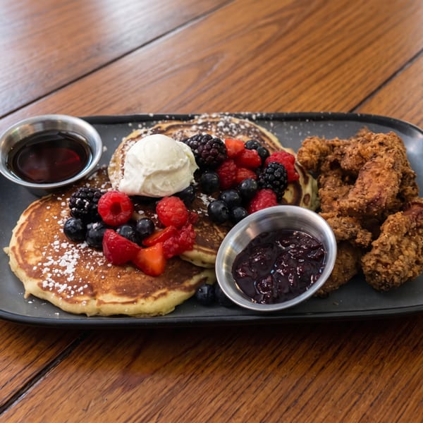 Pancakes with berries, whipped cream, fried chicken, syrup, and jam on a black plate.