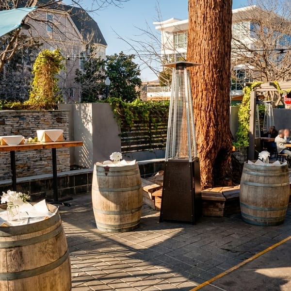 Sunny outdoor restaurant patio with tables, umbrellas, wine barrels, and a stone serving counter.