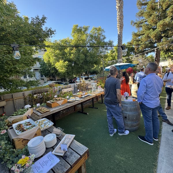 Outdoor gathering with guests mingling around buffet tables filled with food under string lights.