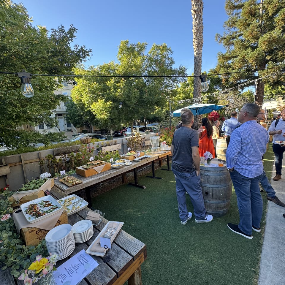 Outdoor gathering with guests mingling around buffet tables filled with food under string lights.