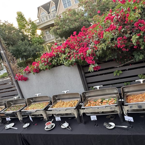 Outdoor buffet line with various dishes on a black table, framed by pink bougainvillea.