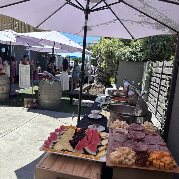 Outdoor buffet table with fruit, cheese, charcuterie, and chafing dishes under white umbrellas at an event.