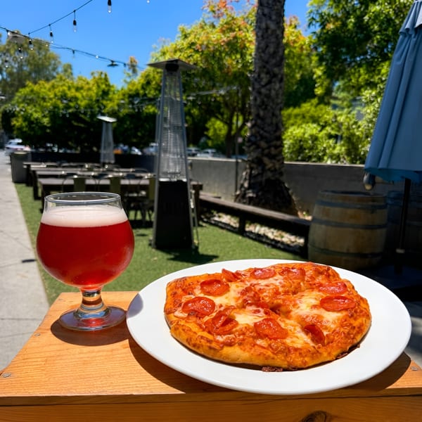 A pepperoni pizza and a red beverage on a wooden table at an outdoor restaurant patio.