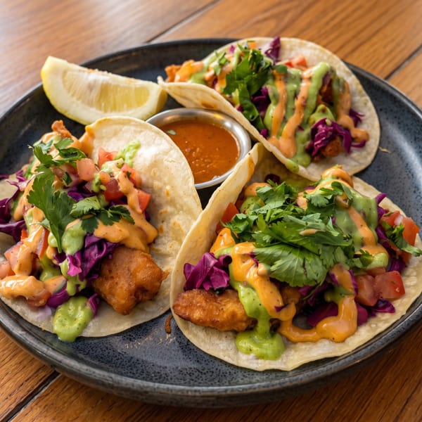 Three vibrant fish tacos topped with purple cabbage, cilantro, and two sauces, plus a lemon.