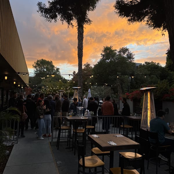 Lively outdoor restaurant patio at dusk with a vibrant sunset, string lights, and many guests.