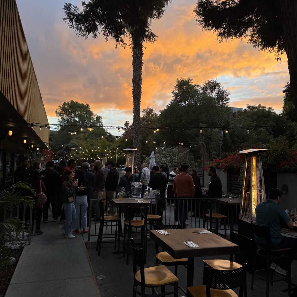 Lively outdoor restaurant patio at dusk with a vibrant sunset, string lights, and many guests.
