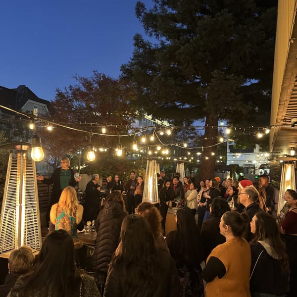 Group of people gathered outdoors at night under string lights and patio heaters, with a woman speaking.